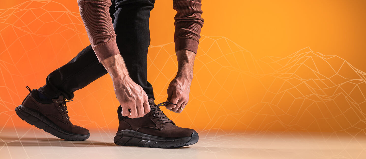 ProTech Verdell para hombre en Lince Dark Brown, mostrado a pie, con las manos de un hombre anudando los cordones del zapato delantero, sobre un fondo degradado naranja/amarillo